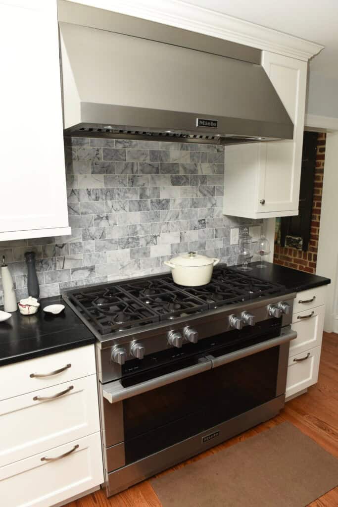 A modern stainless steel gas stove with six burners and a large oven sits between white cabinets in a kitchen. A white Dutch oven is on one burner, and the backsplash features a gray and white marble tile pattern.