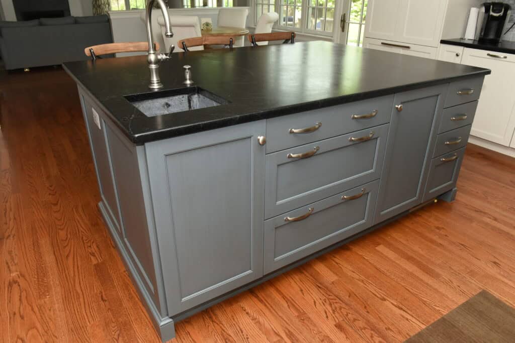 A kitchen island with a dark countertop, built-in sink, silver faucet, and gray cabinets with metal handles, set on a wooden floor in a bright, modern kitchen.