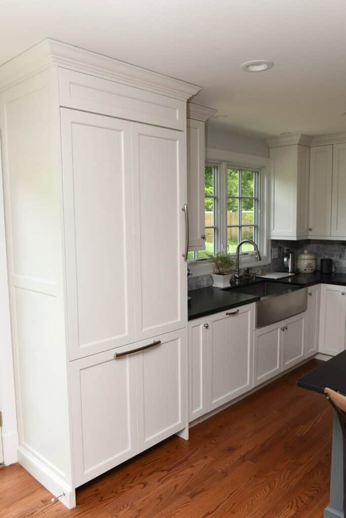 A modern kitchen with white cabinets, stainless steel appliances, a farmhouse sink, black countertops, and large windows letting in natural light. The floor is medium-toned hardwood.