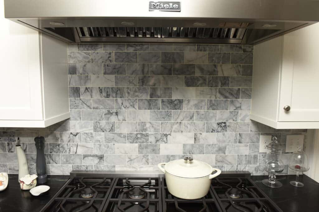 A kitchen stovetop with a white pot on a black gas range, marble tile backsplash, stainless steel vent hood, and countertop items including a pepper grinder, utensils, and glassware.