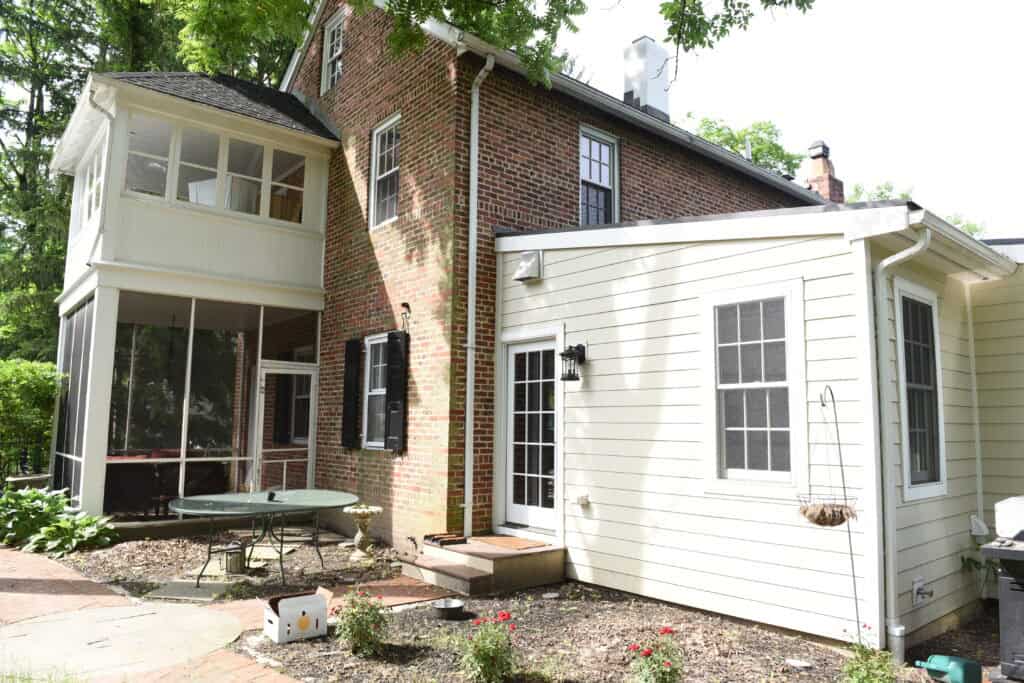 A brick and white-sided house with a screened porch, black shutters, French doors, outdoor table, and hanging planter, surrounded by a small garden and shaded by trees.