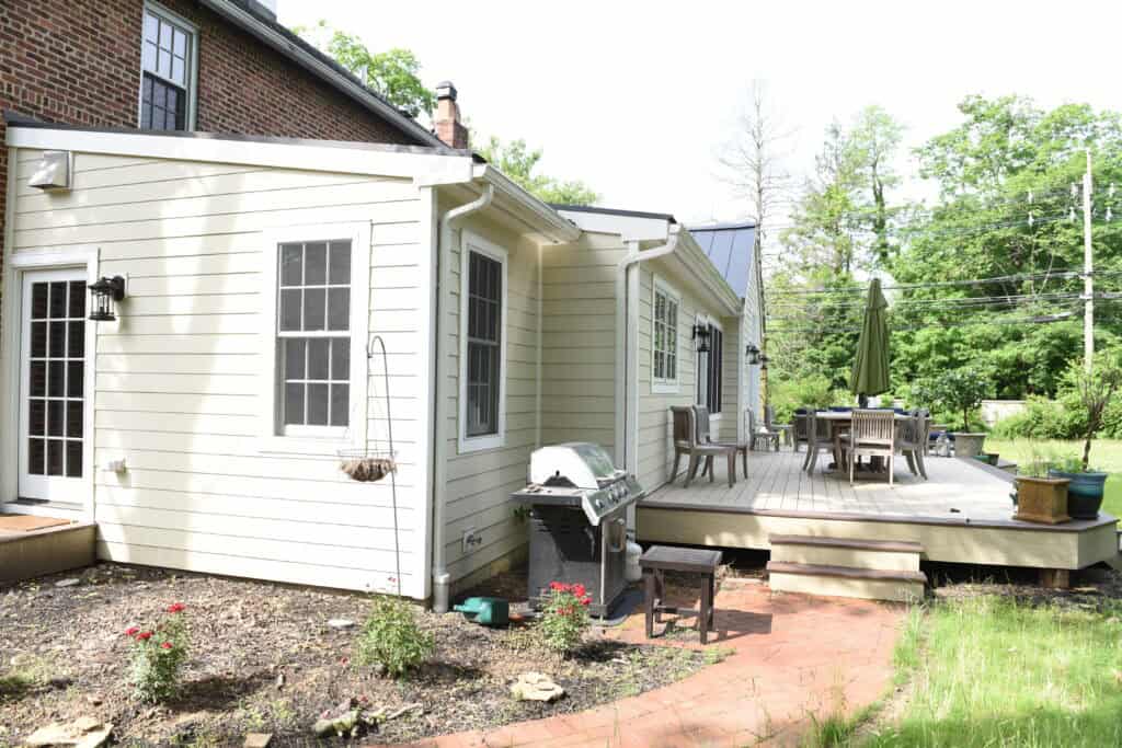 A light-colored house with a small garden, a grill, and a raised wooden deck featuring a dining table with chairs and a green umbrella. The area is surrounded by trees and greenery.
