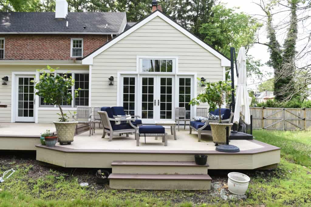 A spacious wooden patio with navy and white outdoor furniture, potted plants, and umbrellas is attached to a cream-colored house with large windows and French doors, surrounded by a grassy yard and trees.