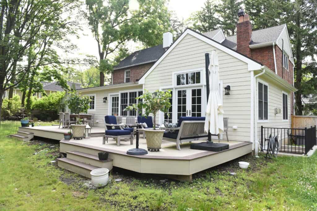 A spacious backyard deck with outdoor seating, potted plants, and umbrellas attached to a light-colored house with large windows and a brick chimney, surrounded by trees and greenery.