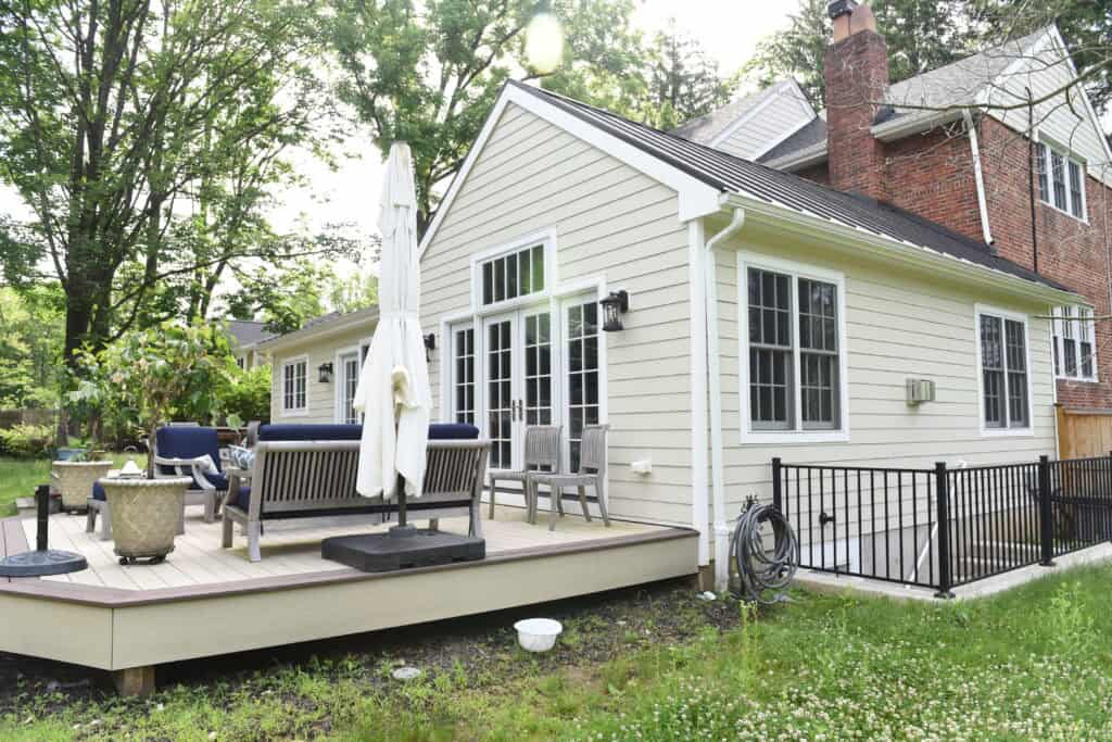 A light-colored house with a small raised wooden deck featuring outdoor furniture, including chairs, a sofa, and a closed umbrella. A garden hose is coiled on the side, and trees surround the backyard.
