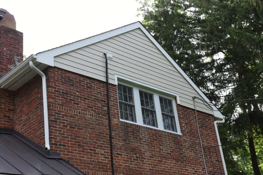 A brick house with white siding on the upper gable, featuring three multi-pane windows and white gutters, surrounded by green trees.
