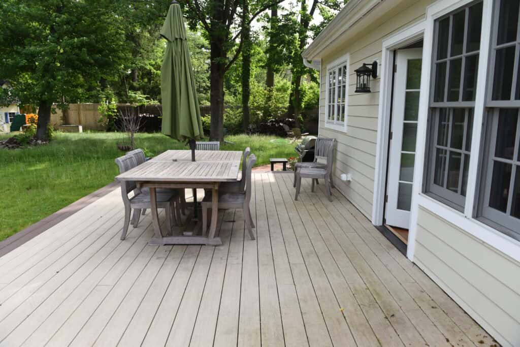 A wooden deck attached to a house with a table, several chairs, and a closed green umbrella. The deck overlooks a grassy backyard with trees and fencing in the background.