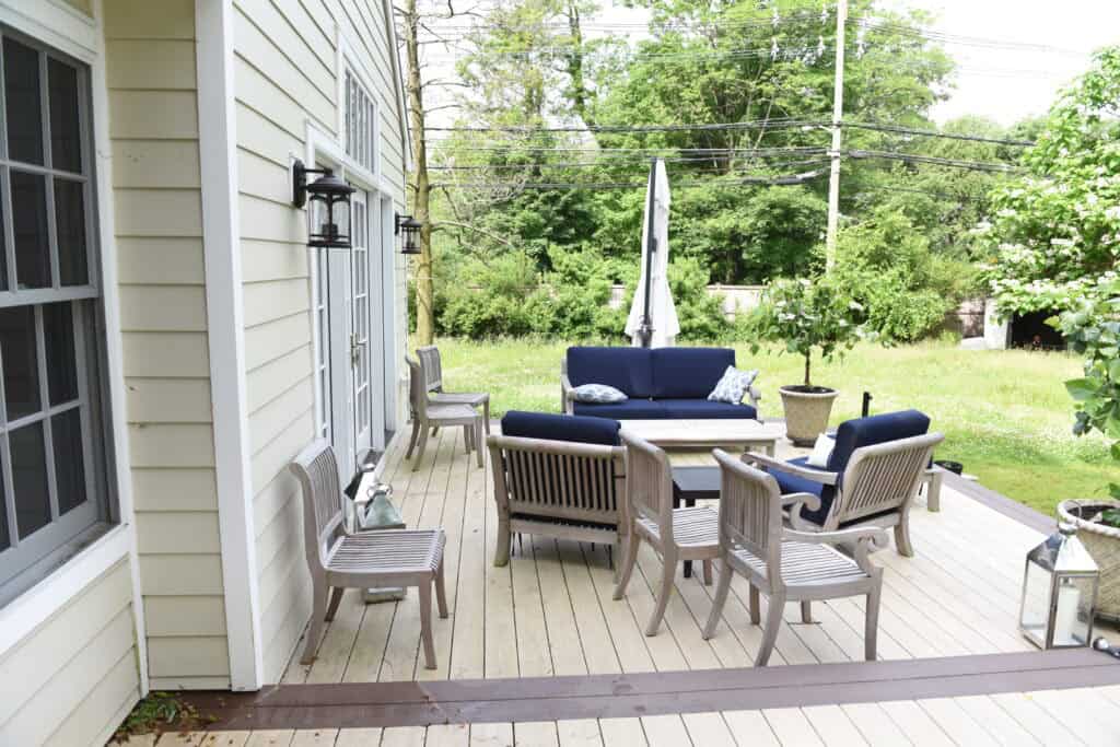 A wooden patio with cushioned seating, wooden chairs, a table, and a closed umbrella. The patio is attached to a cream-colored house and overlooks a grassy backyard with trees and power lines in the background.