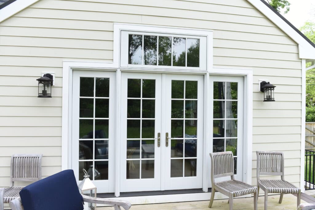 White French doors with window panes lead into a house with beige siding. Two wall-mounted lanterns are on either side of the doors. Wooden chairs and a blue cushion are on the deck in front. Trees are visible in the reflection.