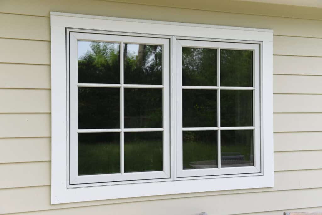 A double-pane window with white trim and grid patterns is set in beige siding. The window reflects green trees and a grassy yard outside.