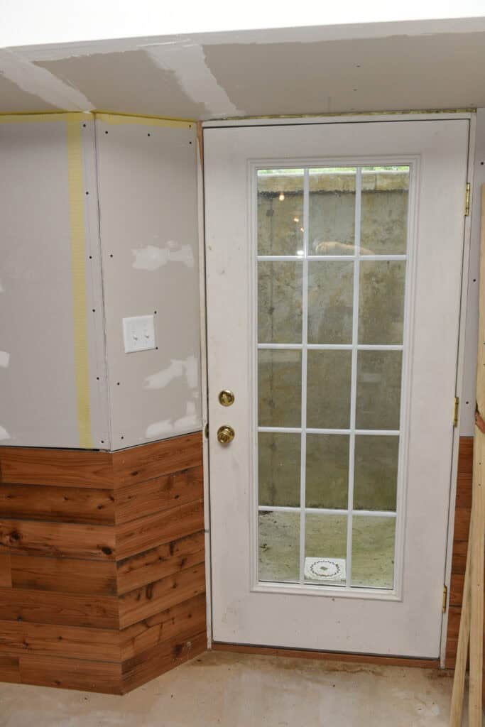 A white door with glass panes leads to a small concrete outdoor space. The interior wall is partially covered with wood paneling and unfinished drywall, with visible screws and patching compound.