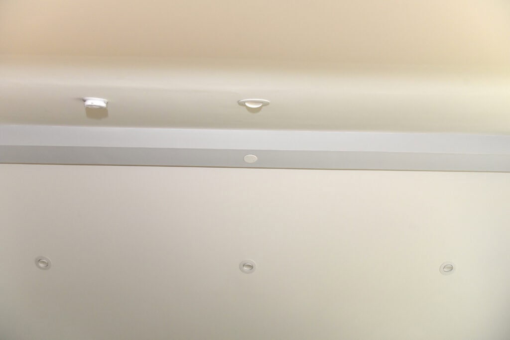 A white ceiling with recessed lighting fixtures and smoke detectors. The ceiling has a slightly raised section with a gray trim running horizontally across the image.