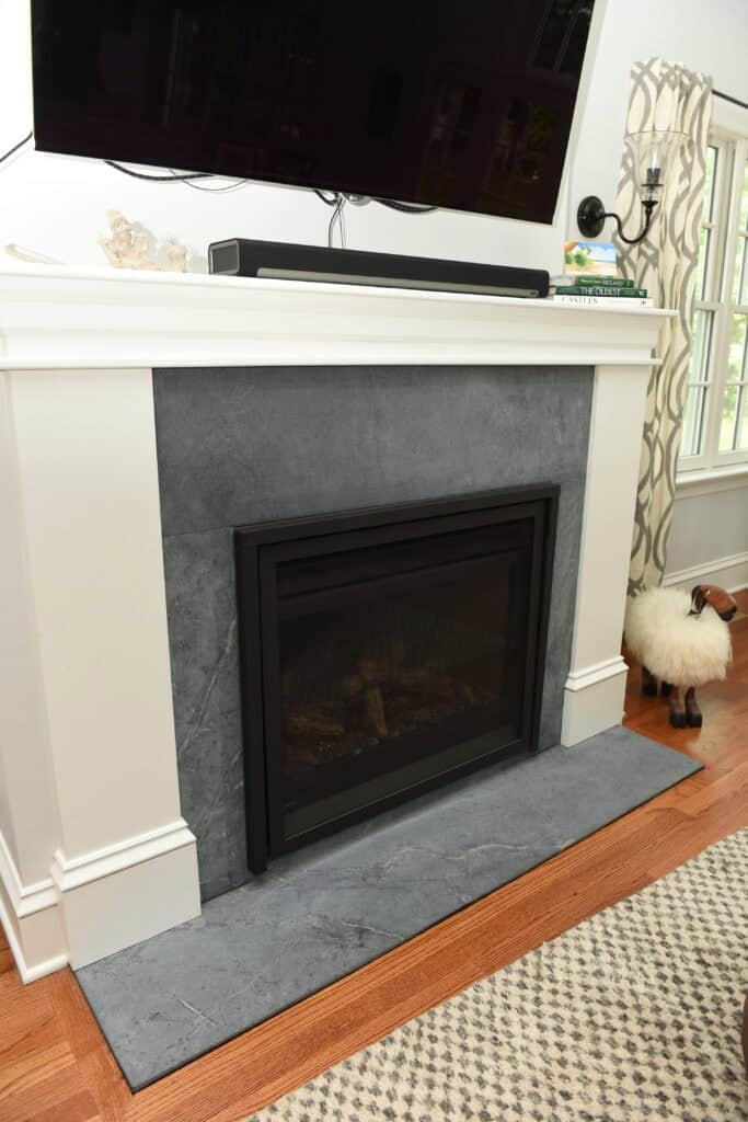 A modern fireplace with a black frame set in a gray stone surround, topped with a white mantel holding decor and books. A TV and soundbar are mounted above. A window with a patterned curtain is to the right.