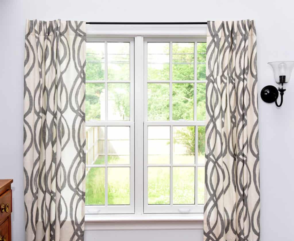 A bright window with white frames and patterned curtains, partially open to reveal a green yard and wooden fence outside. A wall sconce is mounted beside the window.