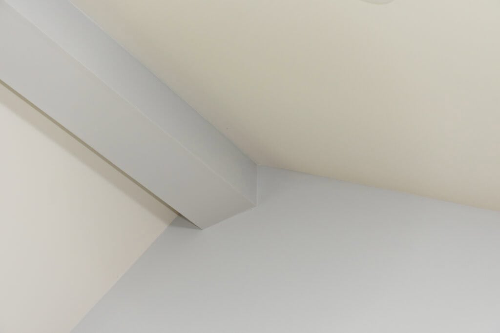 A close-up view of the corner where a ceiling and two walls meet, with a structural beam running diagonally across. The surfaces are painted in light shades of white and gray.