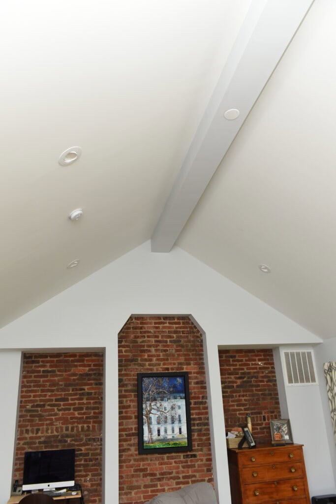 A vaulted white ceiling with recessed lighting above a brick accent wall that features a painting, a computer desk in one alcove, and a wooden dresser in the other.
