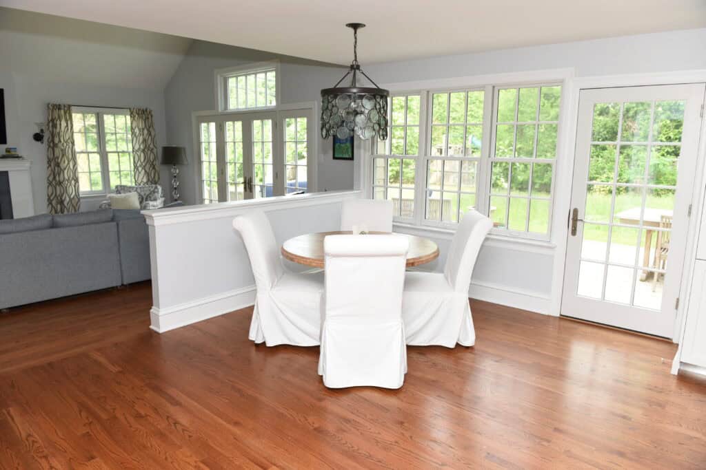 A bright dining area with a round table and four white slipcovered chairs, next to large windows and glass doors overlooking a green outdoor space. Hardwood floors and part of a gray living room sofa are visible.