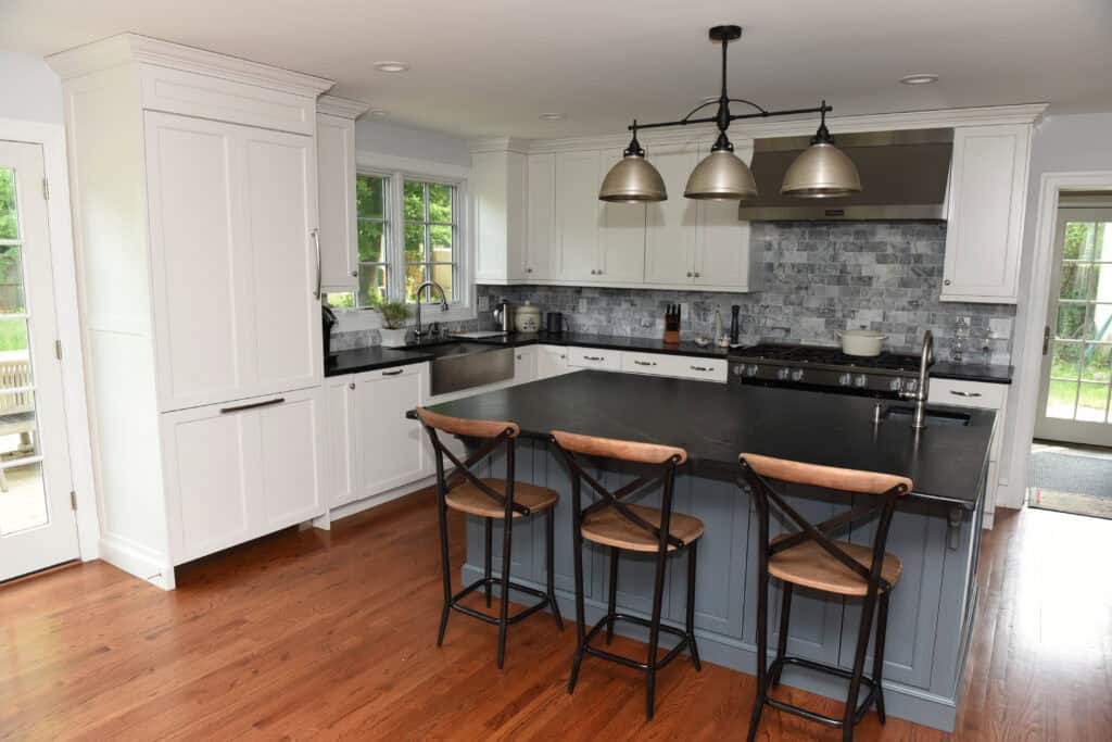 A modern kitchen with white cabinets, a large black island with three wooden stools, pendant lights above, stainless steel appliances, and wood flooring. There are windows and a door letting in natural light.