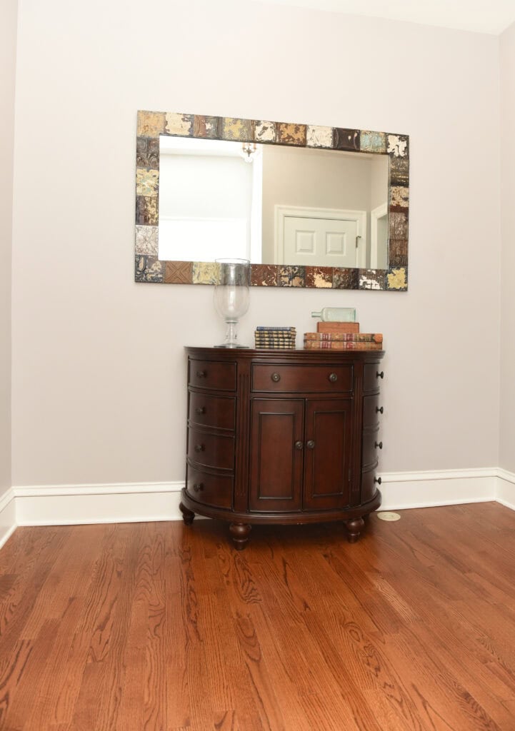 A dark wooden cabinet with books and a glass vase on top sits against a light wall, beneath a large decorative square mirror; the floor is polished wood.