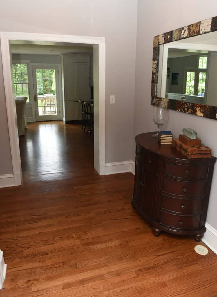 A hallway with polished wooden floors, a dark wood cabinet with books and a glass vase, a large framed mirror above it, and an open doorway leading to a bright room with chairs and large windows.