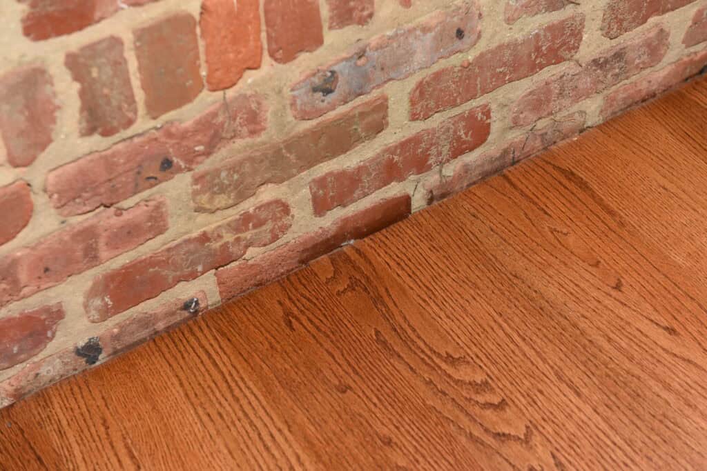 Close-up view of a red brick wall meeting a polished wooden floor with visible wood grain patterns. The bricks are unevenly colored and textured, and the floor appears smooth and well-finished.