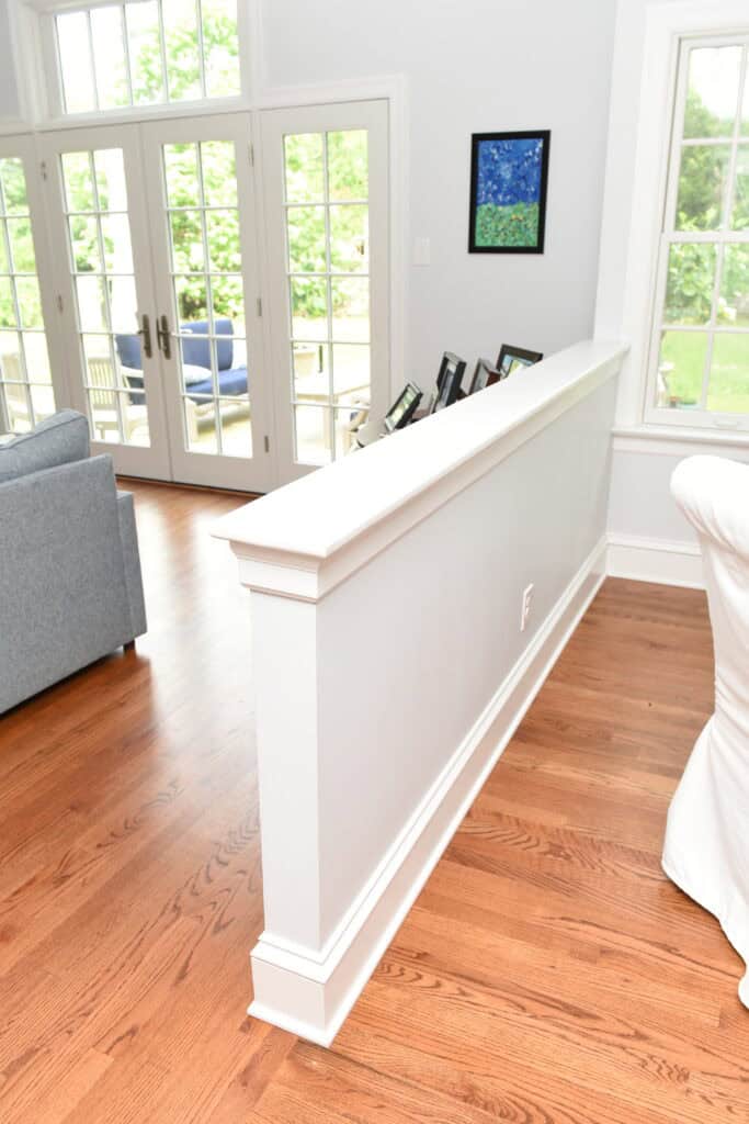 A bright living room with hardwood floors, a gray sofa, a white chair, large windows and glass doors, and a half wall displaying framed photos. Green trees are visible outside.