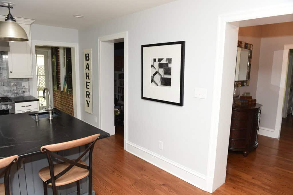 A modern kitchen with black countertops, two wooden bar stools, white cabinets, a BAKERY sign on the wall, and a framed abstract black-and-white artwork in a hallway with hardwood floors.
