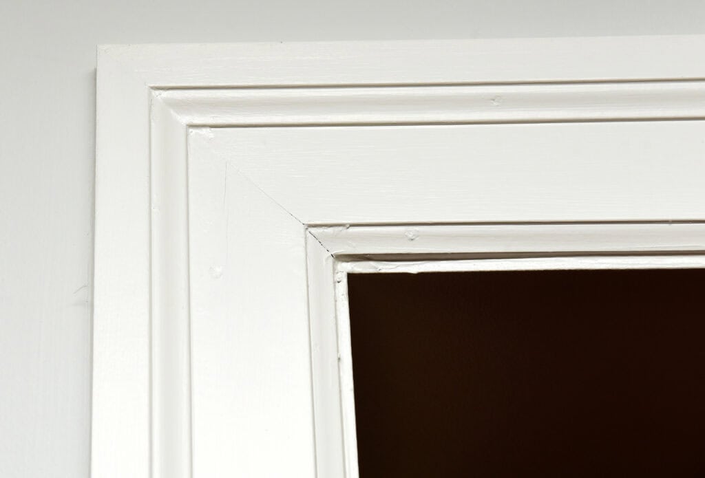 Close-up of the top corner of a white door frame against a white wall, with part of the dark room interior visible through the doorway.