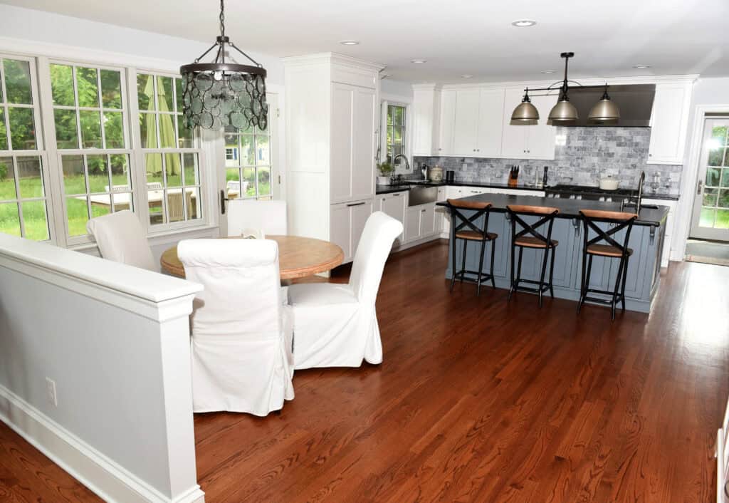 A bright kitchen with hardwood floors, white cabinets, a gray tile backsplash, a round dining table with white slipcovered chairs, and a kitchen island with three black barstools. Large windows look out to a green yard.