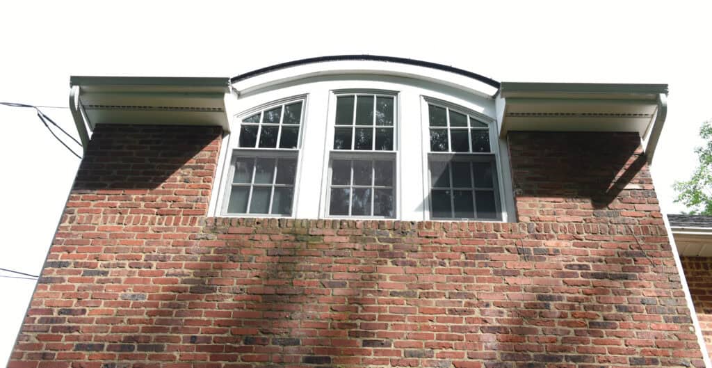 A close-up view of a brick house exterior featuring a large, arched window with multiple panes and white trim, set against a mostly bright sky.
