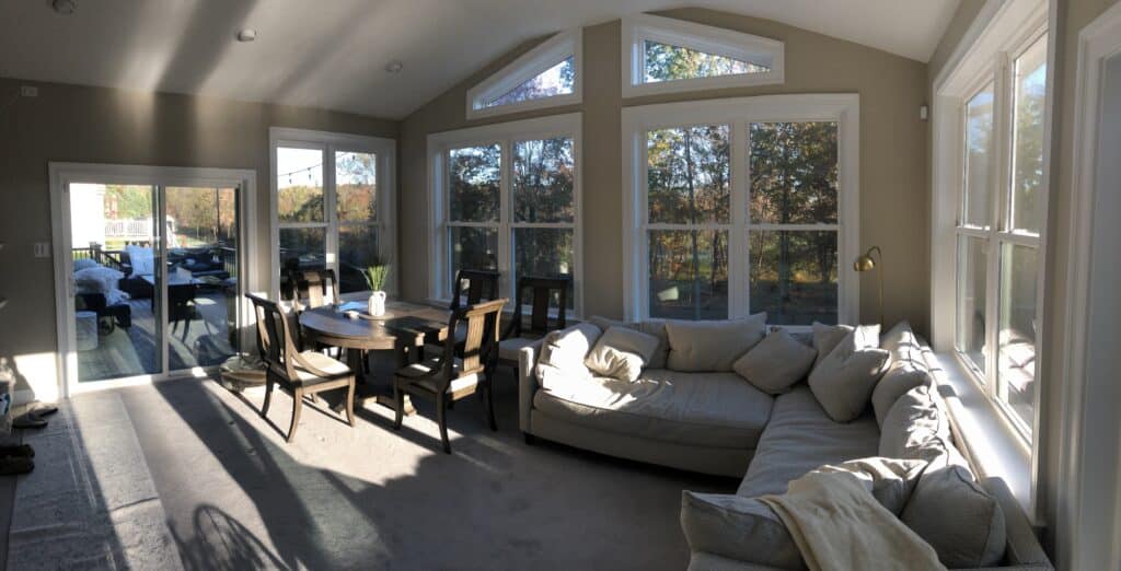 A bright sunroom with large windows, a beige sectional sofa, a round dining table with chairs, and sunlight streaming in, casting shadows on the carpeted floor.