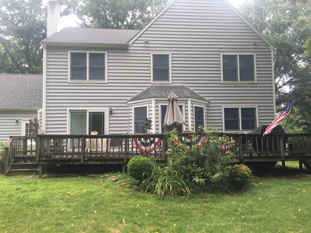 A two-story gray house with white trim, a large wooden deck, patio furniture, and an American flag. The backyard is grassy with some bushes and trees in the background.