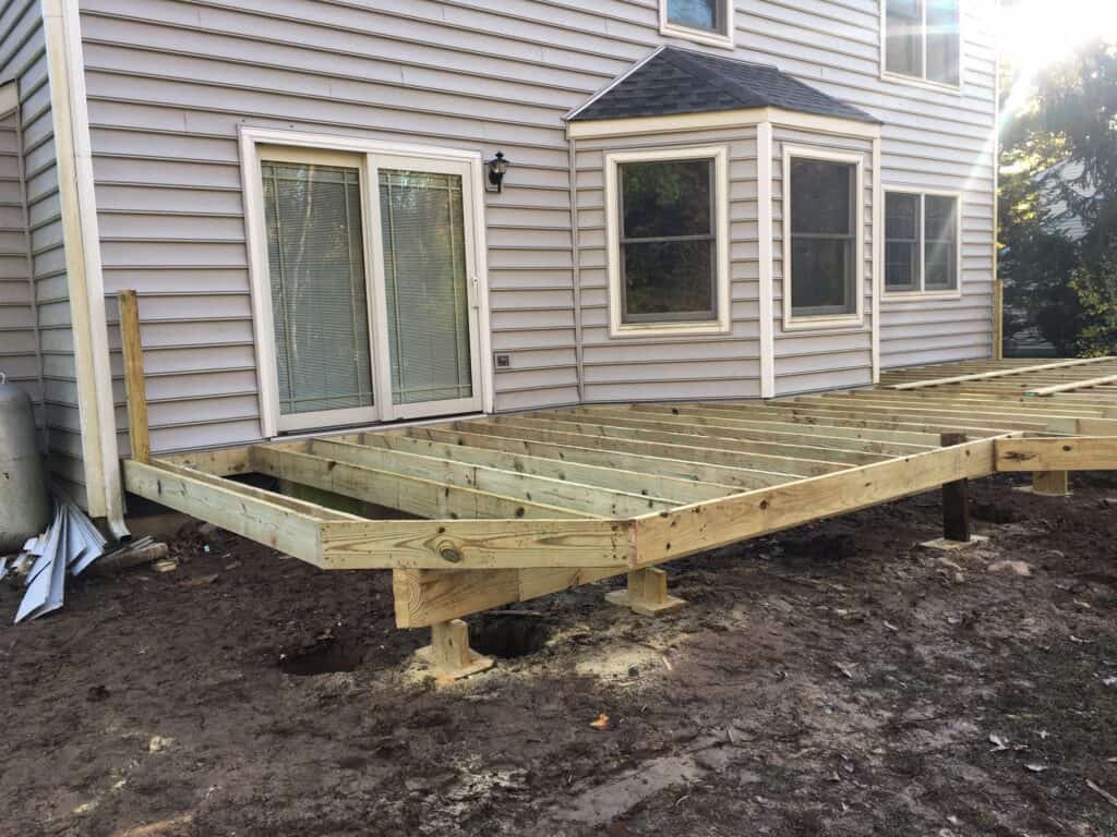 A partially constructed wooden deck is attached to the back of a house with light-colored siding, next to sliding glass doors and beneath bay windows. The ground is bare, and construction materials are visible nearby.