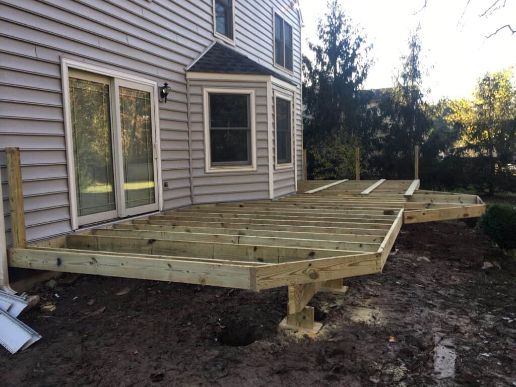 A wooden deck frame is being constructed behind a house, with support posts set in the ground and horizontal beams forming the base structure; the yard is bare with exposed soil.