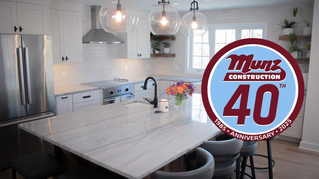 A modern kitchen with white cabinets and a marble island highlights what to expect during a home renovation. The Munz Construction 40th Anniversary logo, in red and blue, is overlaid on the right side, marking 1985–2025.