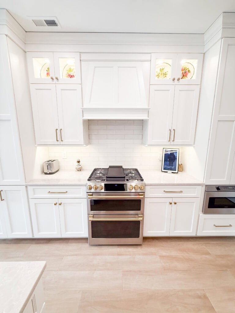 Bright, modern kitchen with white cabinets, gold handles, a stainless steel gas stove, an oven, a toaster, a built-in microwave, a tablet on a stand, and decorative items in lit glass displays above.