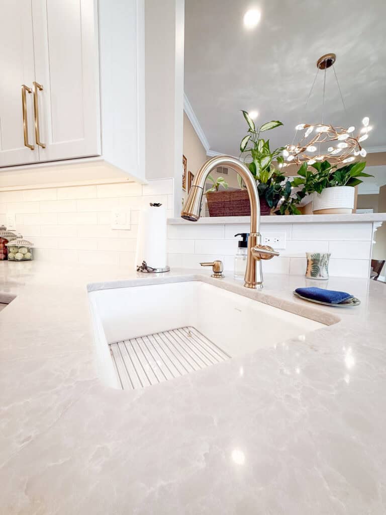 A modern kitchen sink with a gold faucet on a light marble countertop. A paper towel holder, soap dispenser, sponge, and several green potted plants are visible in the bright, clean space.