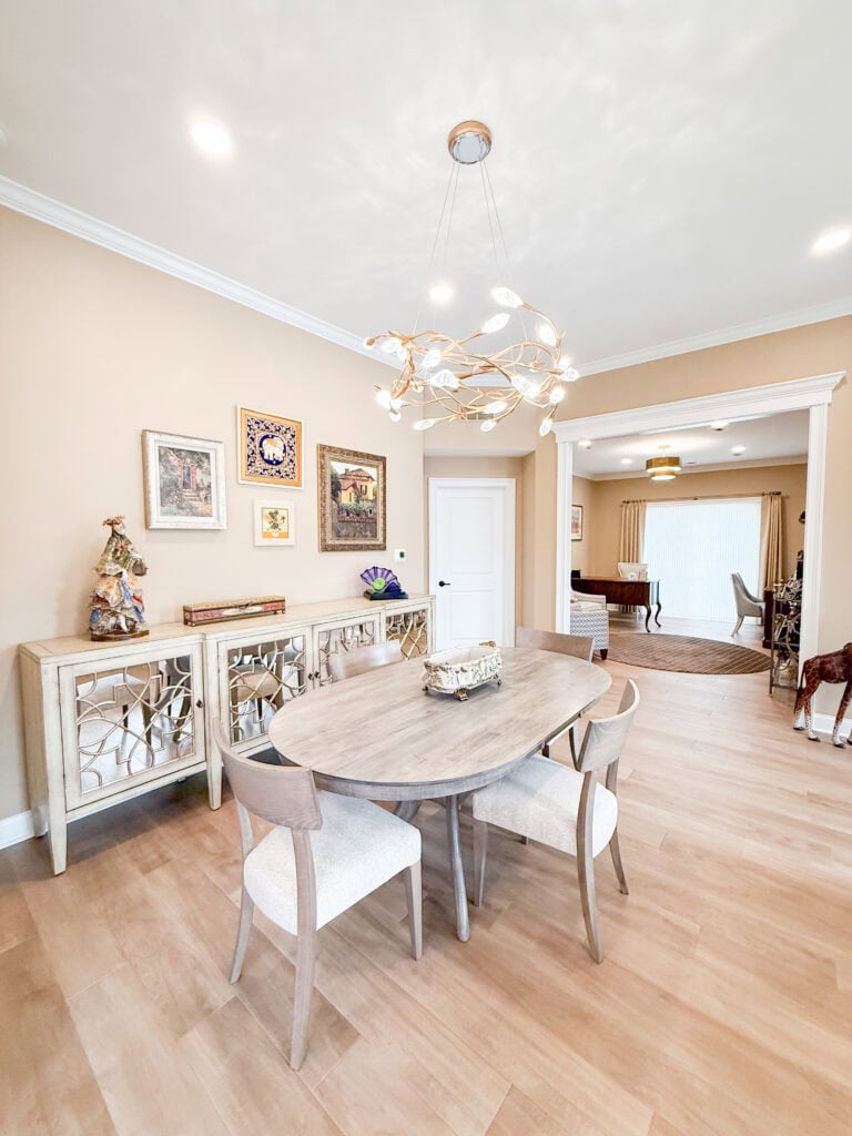 A modern dining room with a light wood oval table and four chairs, a decorative sideboard with artwork above, and a contemporary chandelier. Light wood flooring and neutral walls create a bright, airy atmosphere.