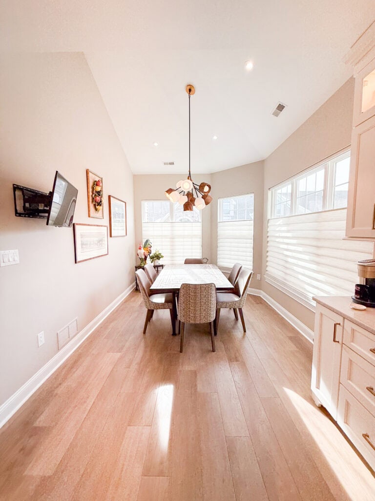 A bright dining room with a rectangular table and six chairs, modern pendant light, large windows with shades, wall art, a wall-mounted TV, and light wood flooring. Natural light fills the space.