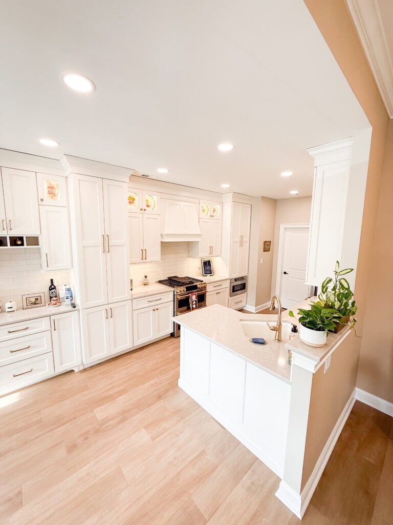 Bright, modern kitchen with white cabinets, gold hardware, stainless steel appliances, light wood flooring, and a white island with a built-in sink and potted plants. Recessed lighting illuminates the space.