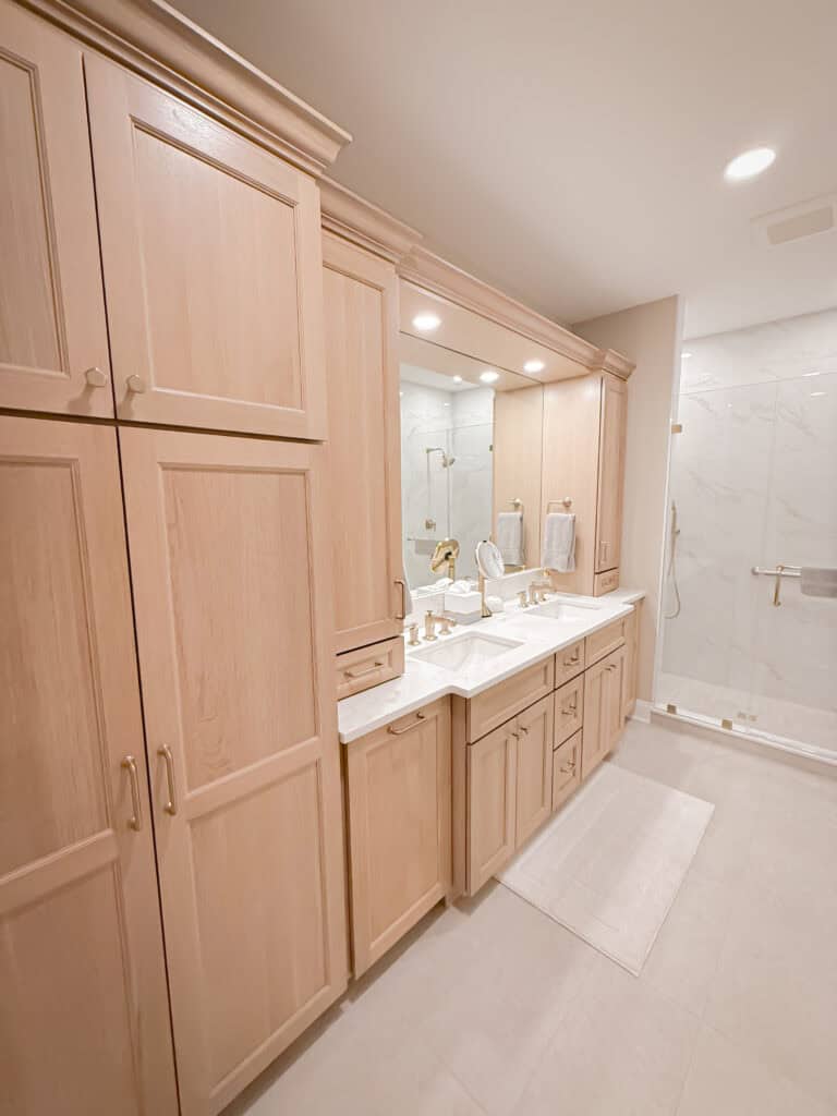 Spacious bathroom with light wood cabinets, dual sinks with gold fixtures, a large mirror, white countertops, and a walk-in shower with glass door and gold accents. Light tiles cover the floor and shower walls.