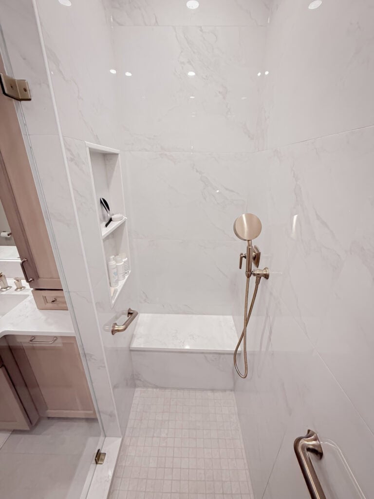 Modern white marble shower with built-in bench, niche shelves holding toiletries, a handheld showerhead, and glass door. A bathroom vanity is partially visible through the doorway.
