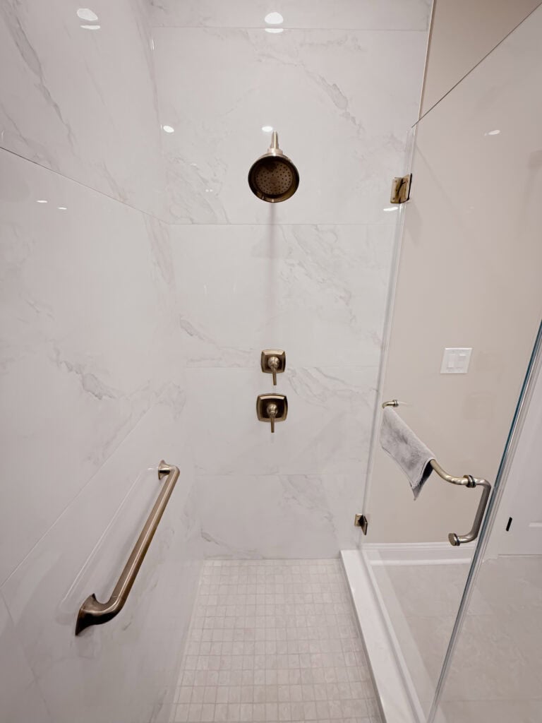 A modern walk-in shower with white marble-look tiles, a rainfall showerhead, gold fixtures, a safety grab bar on the left wall, and a glass door with a towel hanging on a bar.