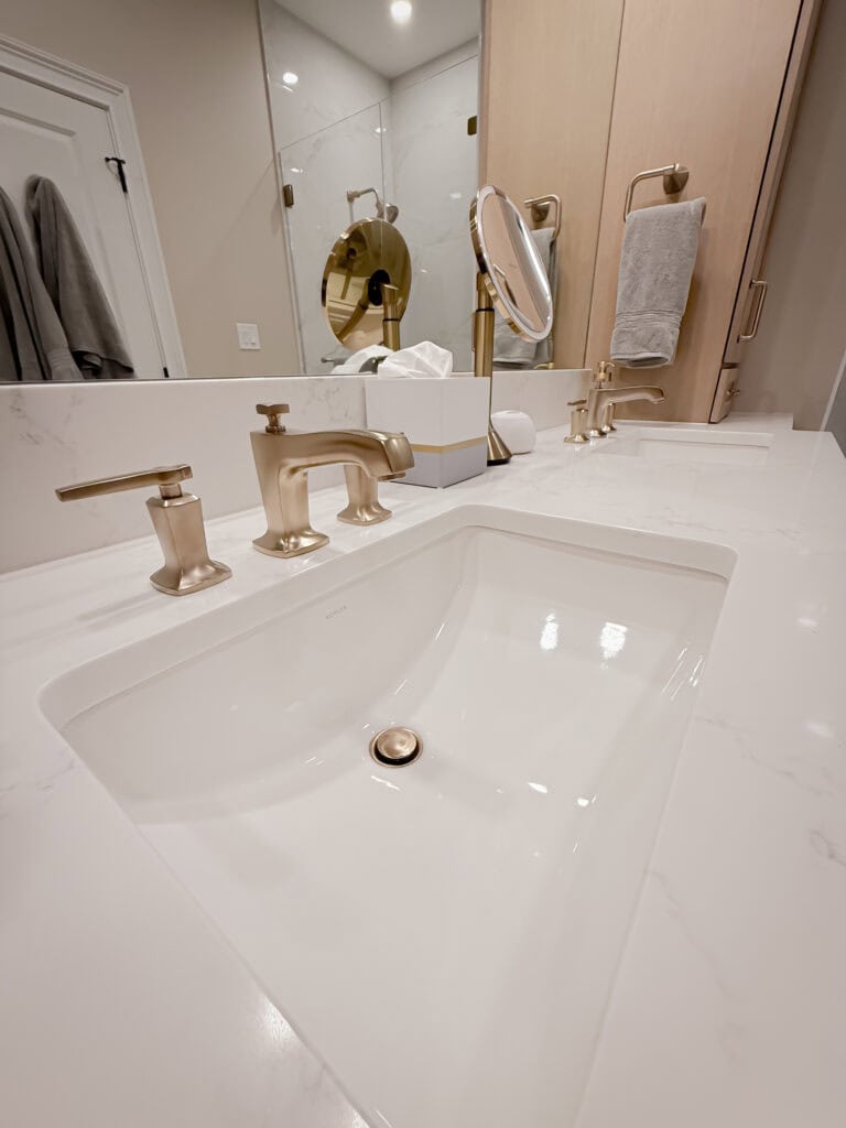 Modern bathroom sink with gold faucet and fixtures, a marble countertop, mirror, towel ring with towel, and minimalist decor. A round magnifying mirror and tissue box also sit on the counter.