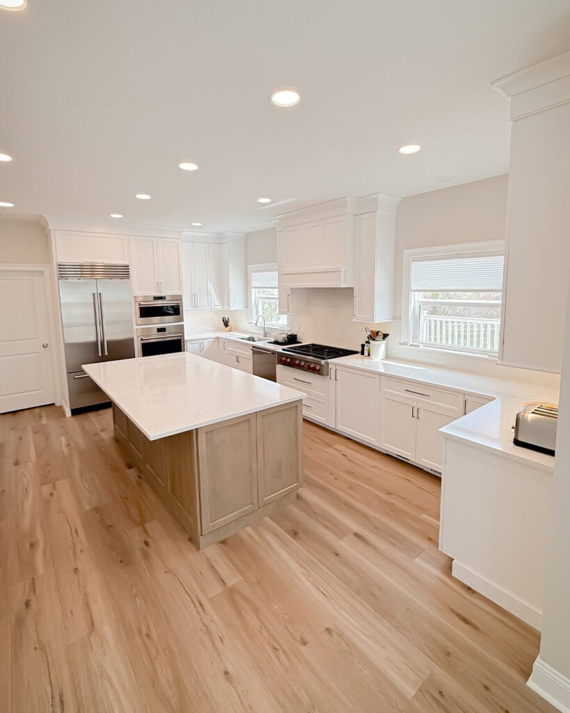 Bright, modern kitchen with white cabinets, stainless steel appliances, an island with a light wood base and white countertop, and light wood flooring. Large windows let in natural light.