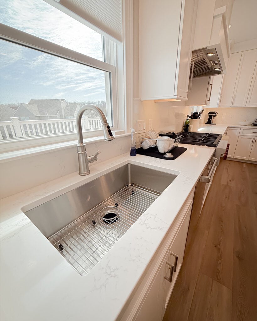 A modern kitchen with a large stainless steel sink under a window, white marble countertops, white cabinets, and a stove with kitchen appliances along the right side. Natural light fills the room.