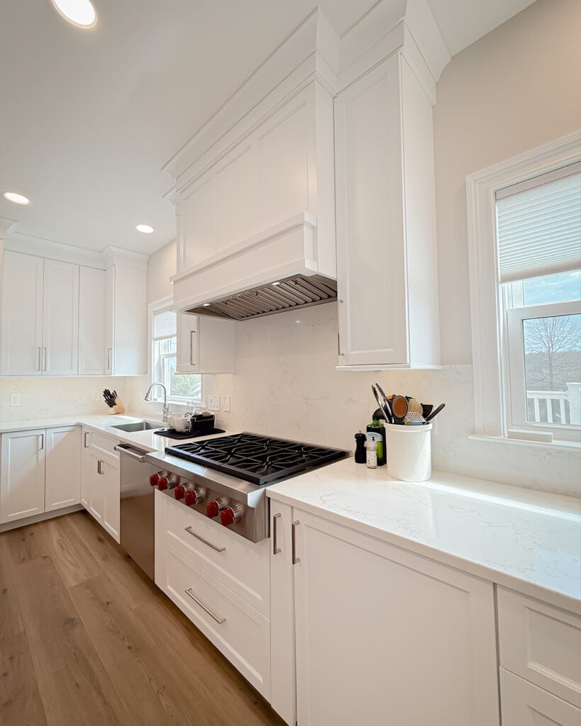 Bright, modern kitchen with white cabinets, marble countertops, a stainless steel gas stove with red knobs, and a pot on the burner. Cooking utensils sit in a white container by the window.