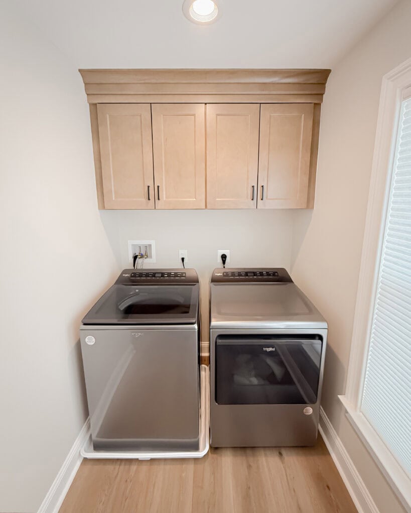A modern laundry room with a stainless steel top-load washer and front-load dryer side by side, light wood cabinets above, neutral walls, and natural light from a window on the right.