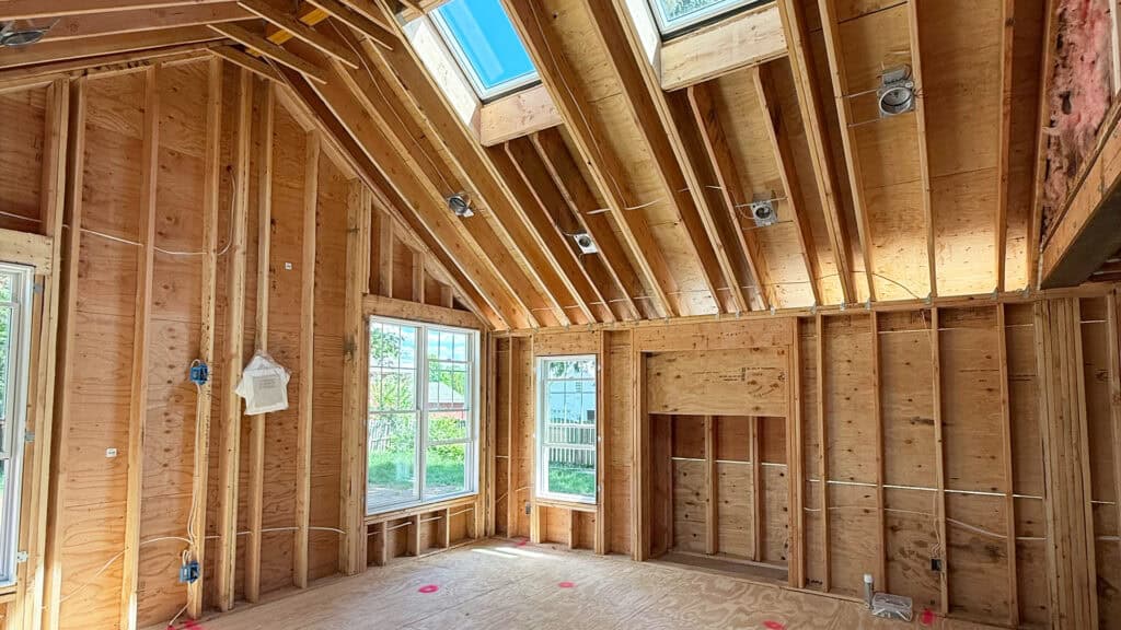 Interior of a house under construction with exposed wooden framing, large windows letting in sunlight, vaulted ceiling with skylights, and visible electrical wiring. No drywall or insulation is installed yet.