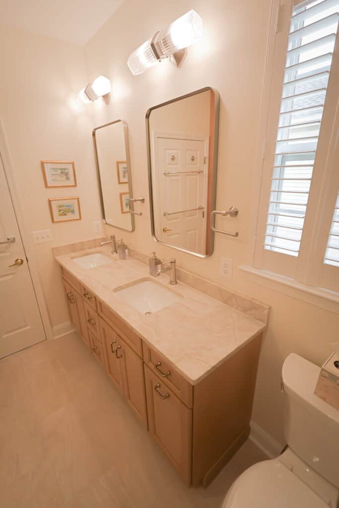 a bright bathroom with a double vanity, two mirrors, marble countertop, dual sinks, modern fixtures, wall lights above each mirror, framed art on the wall, and a window with white shutters.